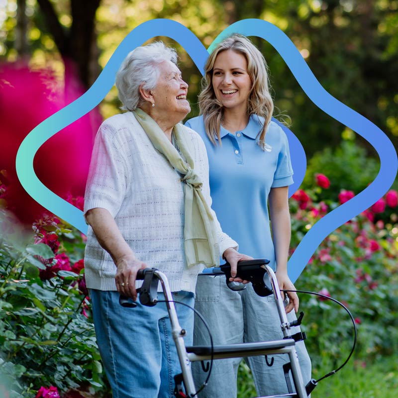 A carer walks with an older woman through a rose garden